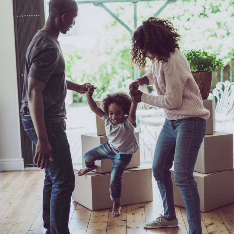 A photo of playful parents holding sons's hands in new house. Happy and playful family are with cardboard boxes. They are in casuals.