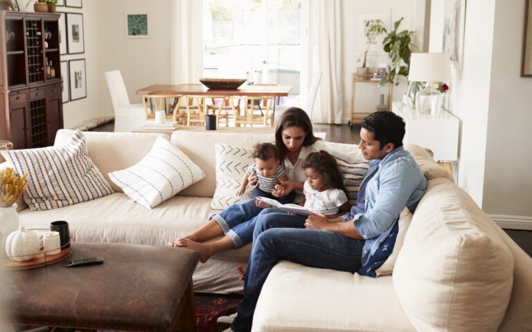 Young Hispanic family sitting on sofa reading a book together in the living room, seen from doorway